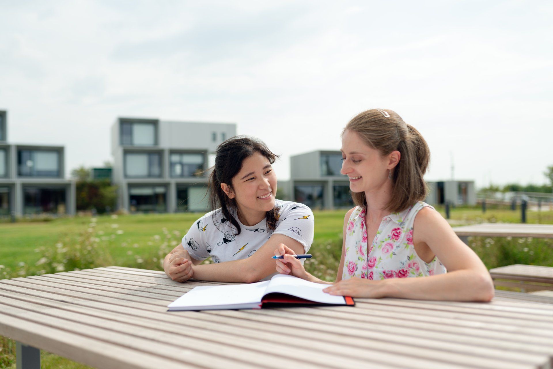 Two people reading a book together at a picnic table outdoors on a sunny day