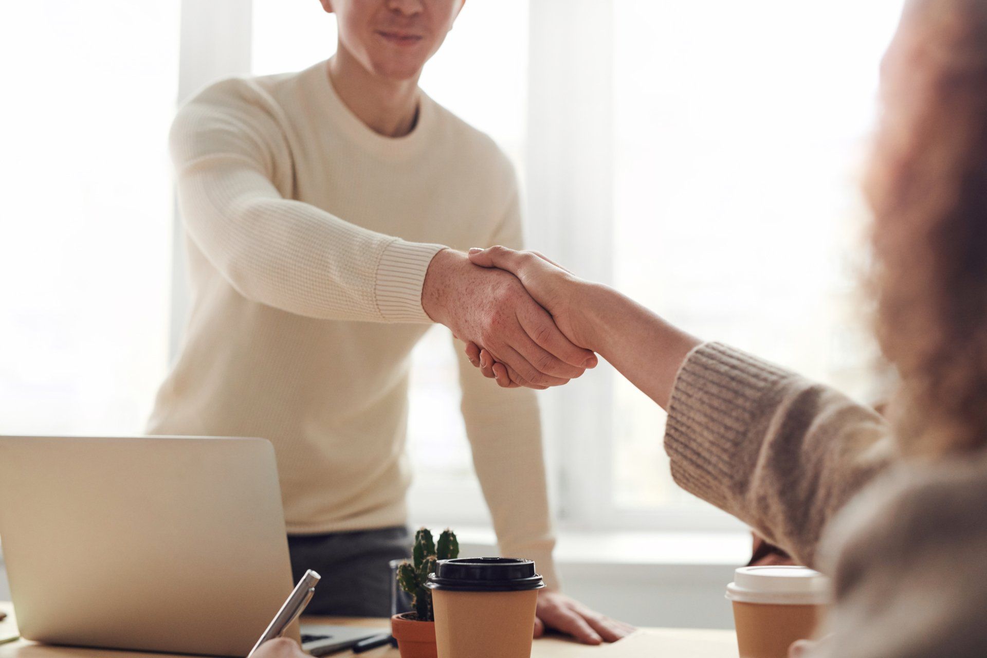 Two people shaking hands across a desk with a laptop and coffee cups in a bright office