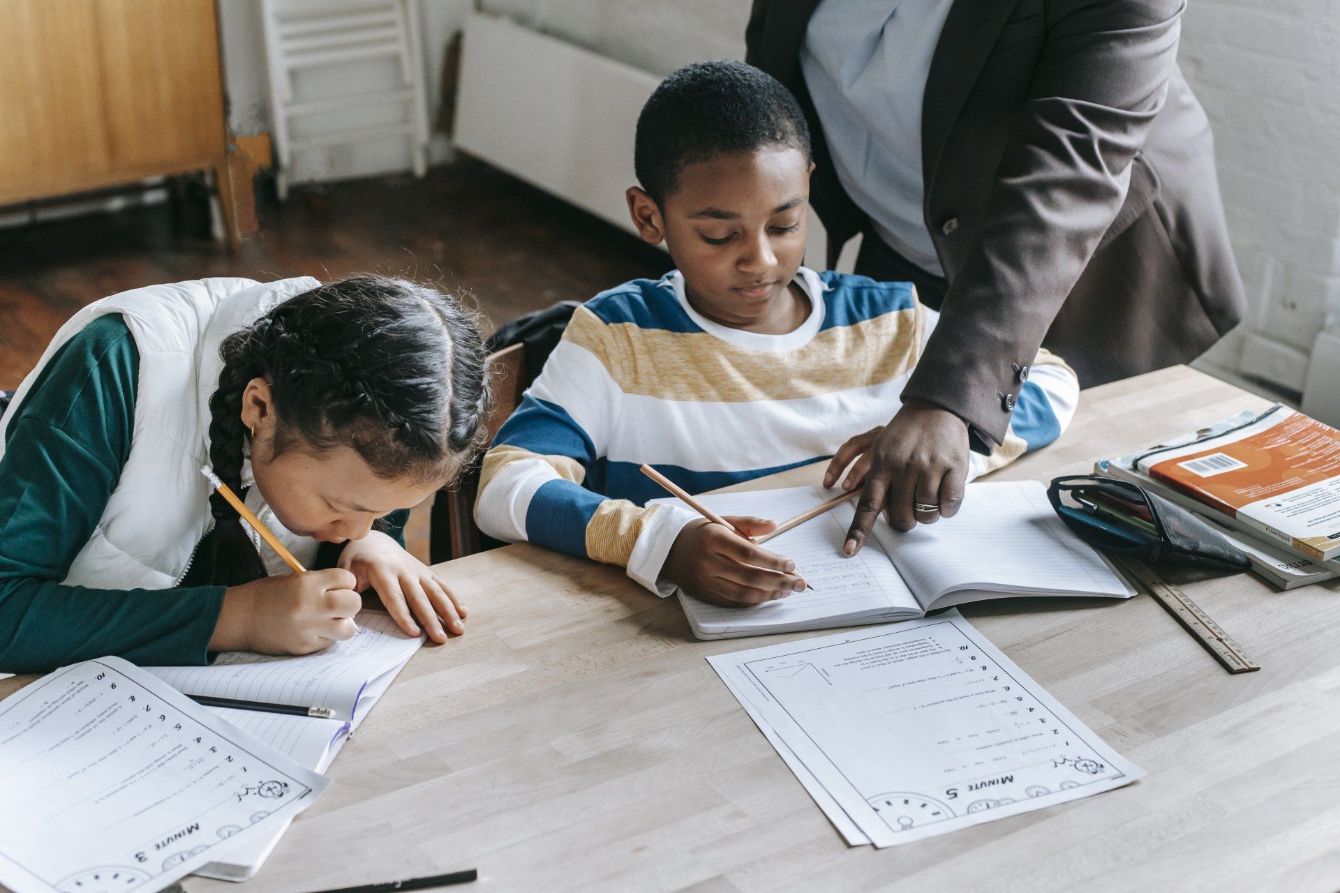 Two children study at a table while an adult points to a notebook.
