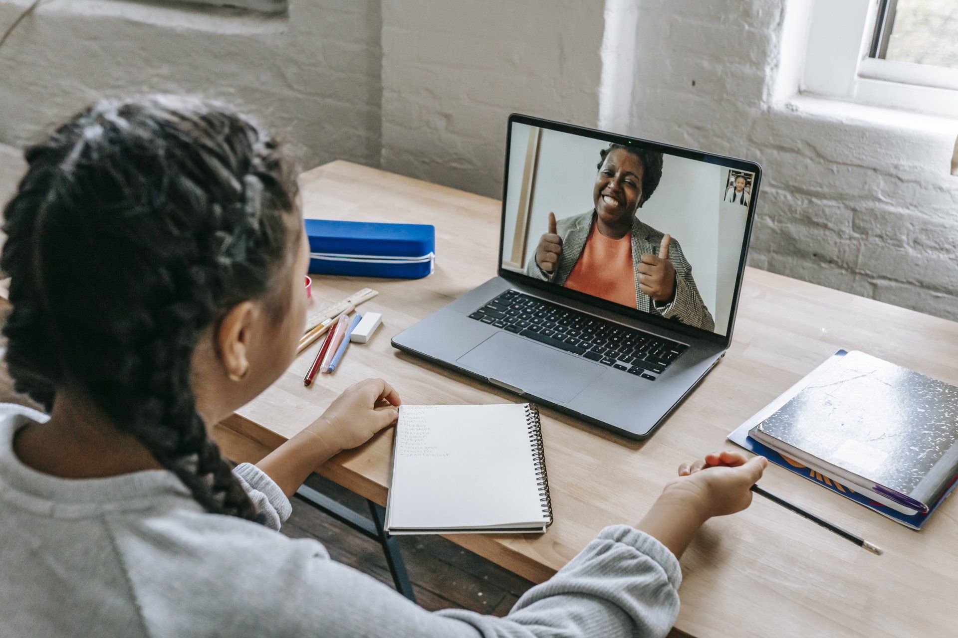 Person taking notes during a video call on a laptop at a desk with books and a notebook