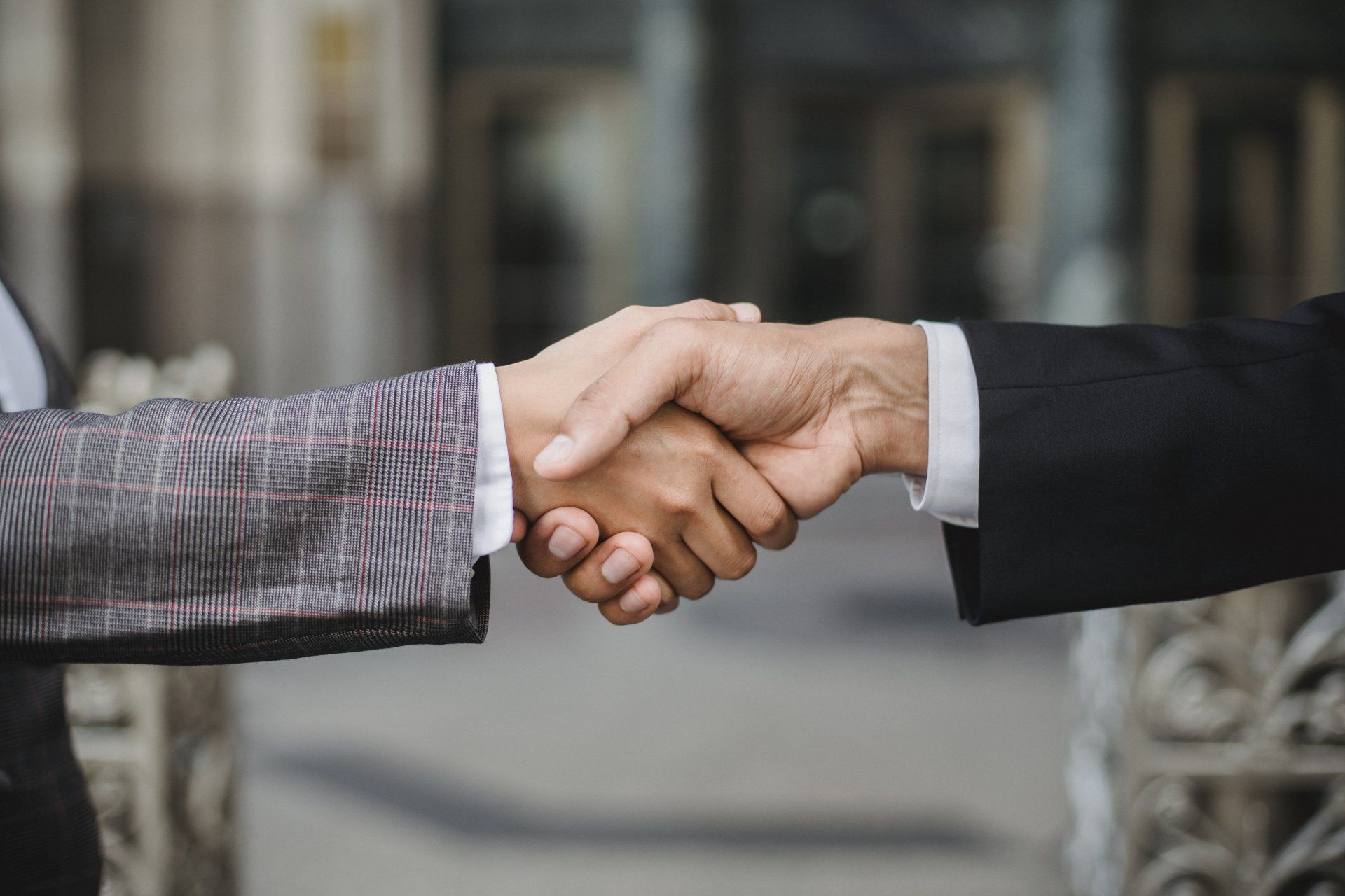 Two people in suits shaking hands outdoors in front of a blurred building.