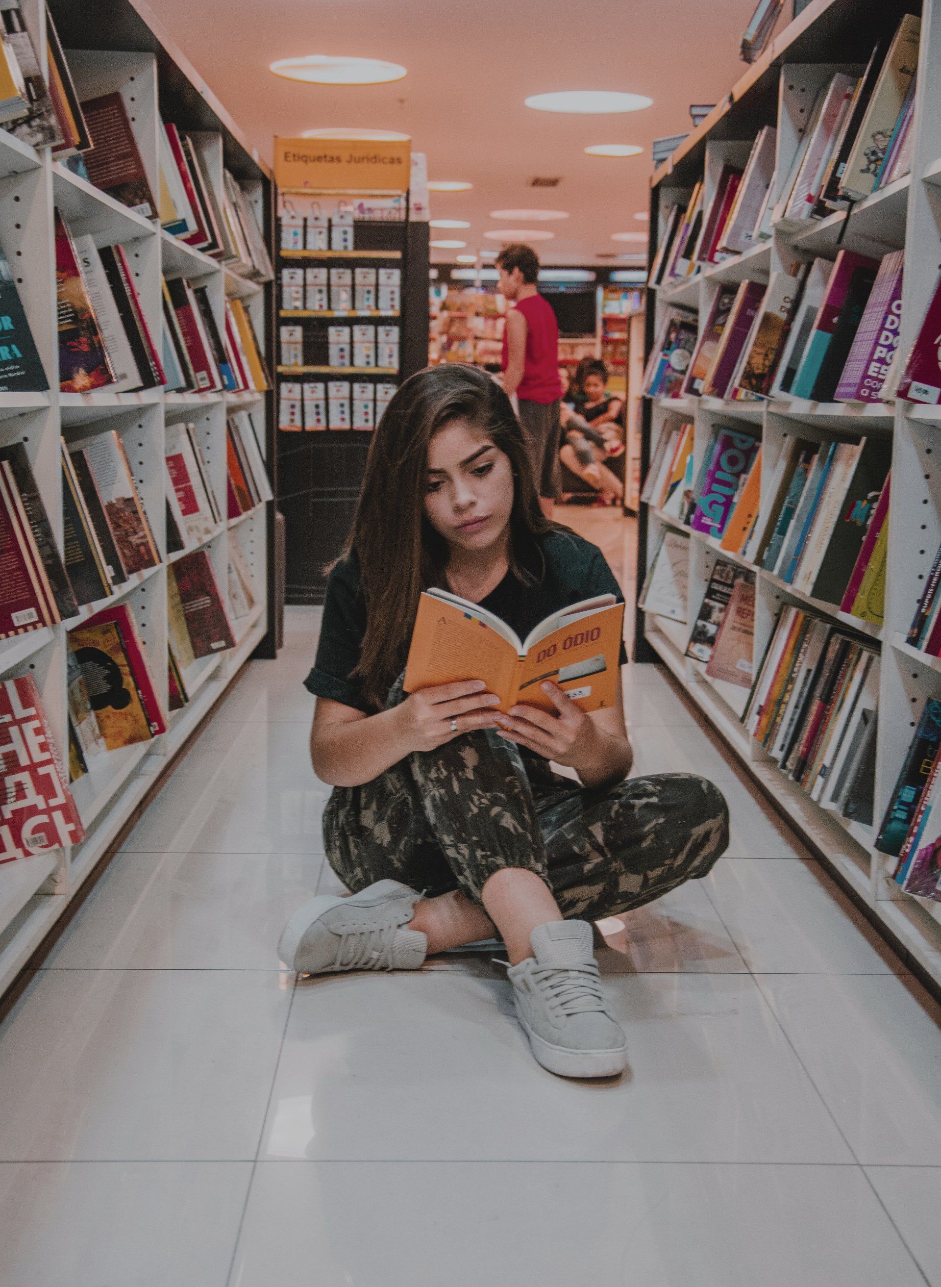 Woman sitting on a library floor reading an orange book between rows of shelves